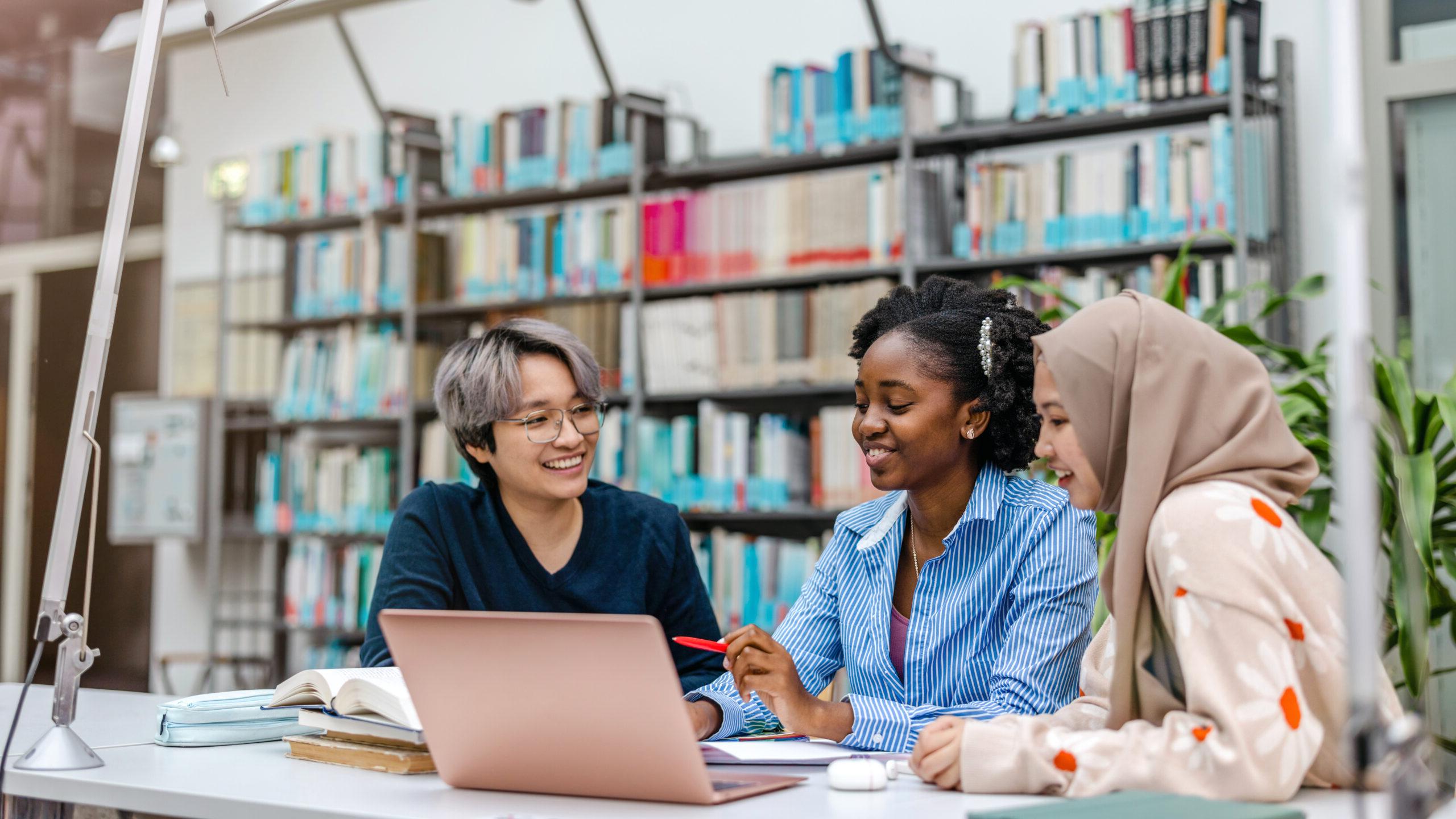 A group of students sitting at a table in a library, smiling and working together on a project with a laptop and books.